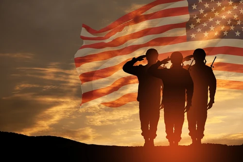 Silhouettes of three soldiers saluting against a sunset sky with an American flag overlay.